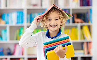 Lachender Schüler mit bunten Büchern auf dem Kopf in der OGS.